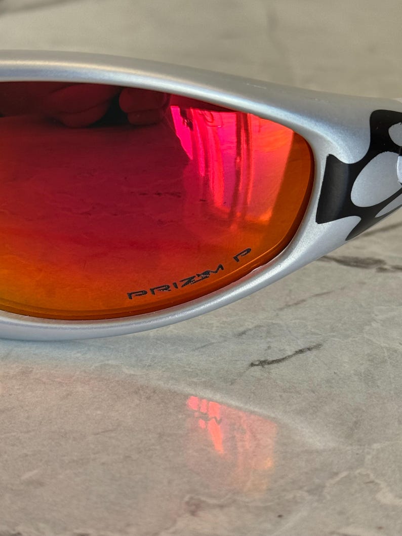 May include: Silver-framed sunglasses with red-orange lenses. The lenses have the text "PRIZM P" printed on them. The frame has a black and white design on the temple.