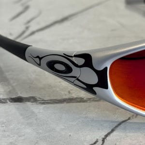 May include: Silver sunglasses with a black and gray tribal design on the frame. The lenses are a vibrant red-orange color. The sunglasses are angled, showing the details of the frame and lens.