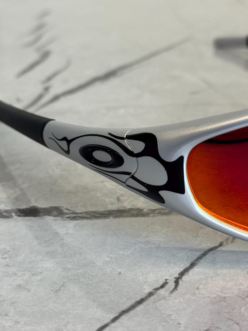 May include: Silver sunglasses with a black and gray tribal design on the frame. The lenses are a vibrant red-orange color. The sunglasses are angled, showing the details of the frame and lens.