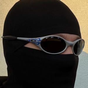 Puede incluir: Gafas de sol plateadas con lentes oscuras y detalles de llamas azules. Las gafas de sol se llevan con un pasamonta&ntilde;as negro. La imagen est&aacute; tomada con luz natural, con un fondo neutro.