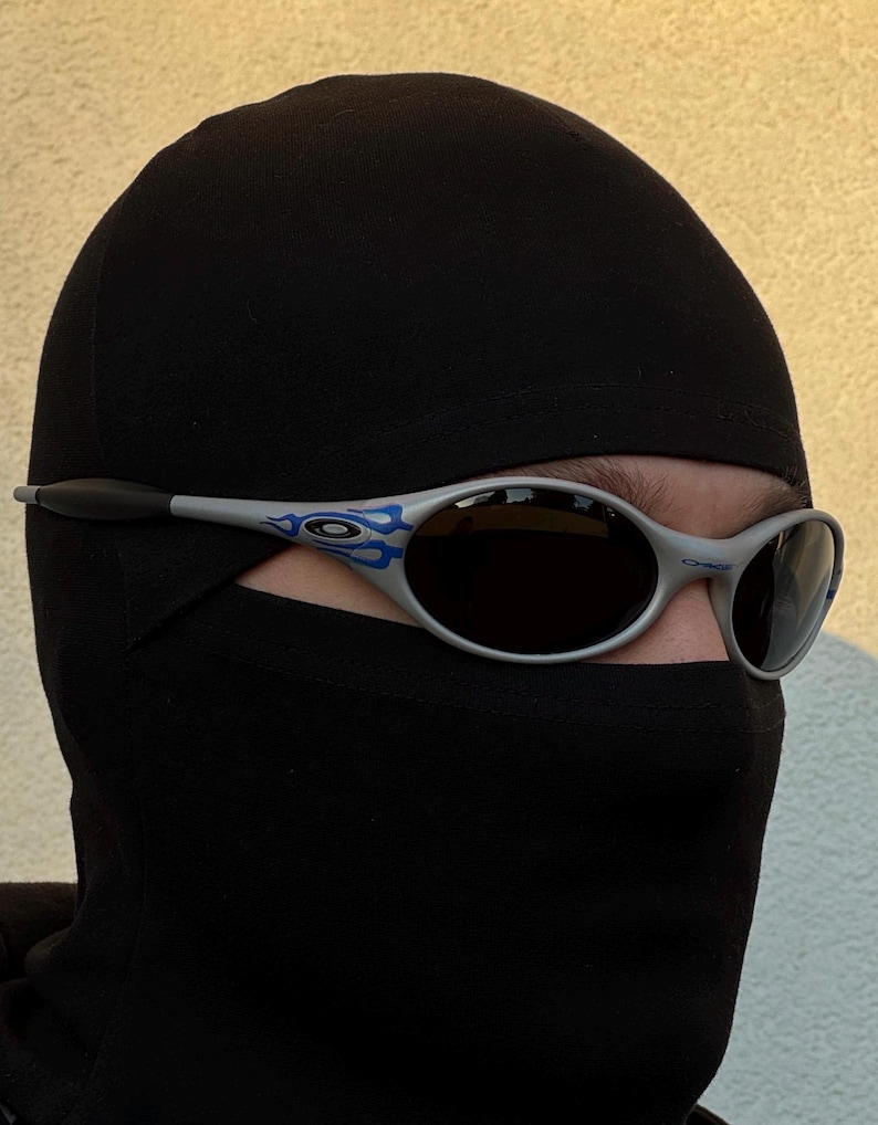 Puede incluir: Gafas de sol plateadas con lentes oscuras y detalles de llamas azules. Las gafas de sol se llevan con un pasamonta&ntilde;as negro. La imagen est&aacute; tomada con luz natural, con un fondo neutro.