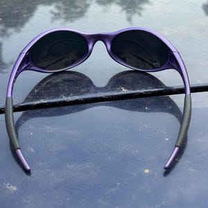 May include: Purple sunglasses with black lenses. The sunglasses have a sleek, modern design with a curved frame and dark lenses. The arms of the sunglasses are black at the ends. The sunglasses are resting on a dark blue surface.