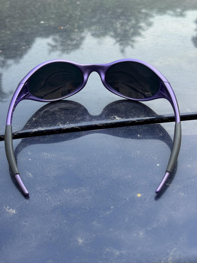May include: Purple sunglasses with black lenses. The sunglasses have a sleek, modern design with a curved frame and dark lenses. The arms of the sunglasses are black at the ends. The sunglasses are resting on a dark blue surface.