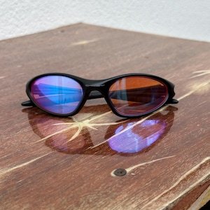 May include: Black sunglasses with mirrored lenses. The lenses reflect blue and orange hues. The frame is black and has a sleek, curved design. The sunglasses are resting on a wooden surface.