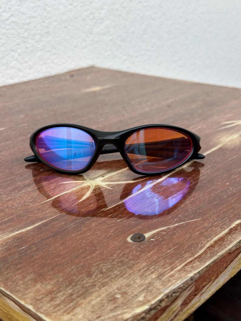 May include: Black sunglasses with mirrored lenses. The lenses reflect blue and orange hues. The frame is black and has a sleek, curved design. The sunglasses are resting on a wooden surface.