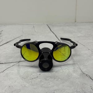 May include: Black sunglasses with yellow lenses. The glasses have a unique design with a central nose piece and a curved frame. The lenses reflect a yellow hue, and the frame appears to be made of a durable material.