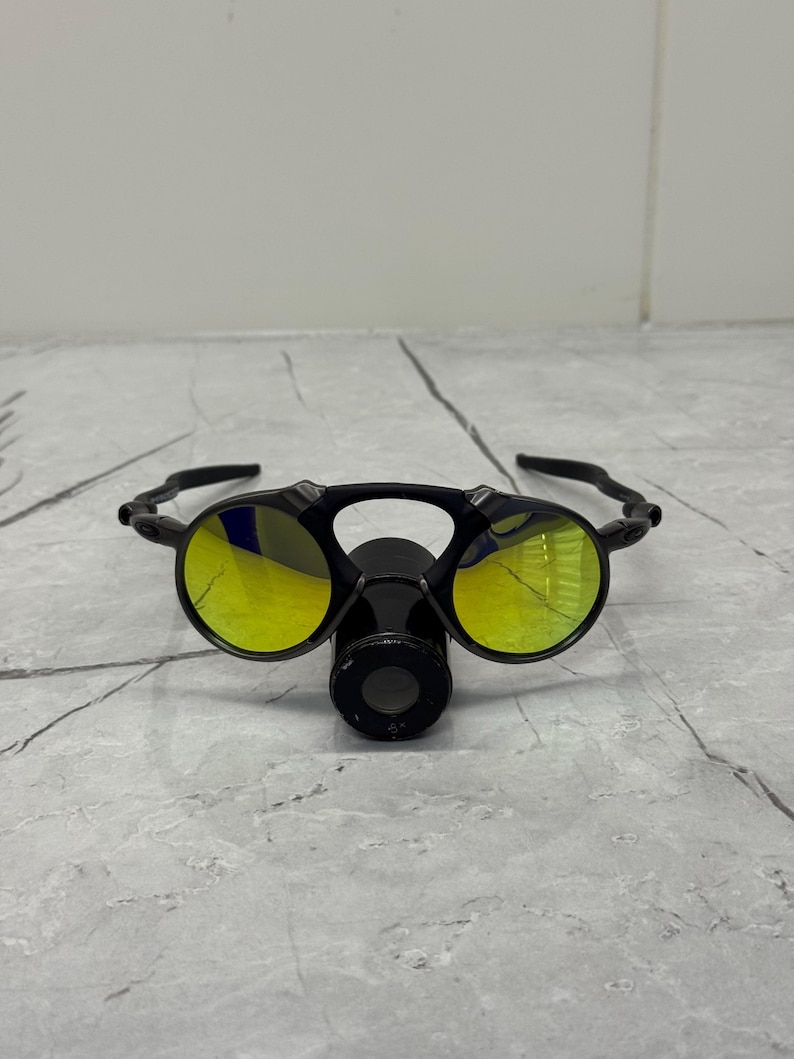 May include: Black sunglasses with yellow lenses. The glasses have a unique design with a central nose piece and a curved frame. The lenses reflect a yellow hue, and the frame appears to be made of a durable material.