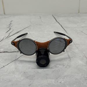 May include: Round sunglasses with dark lenses and a brown leather bridge. The frame features black arms and decorative stitching on the leather. A black cylindrical object is positioned below the glasses.