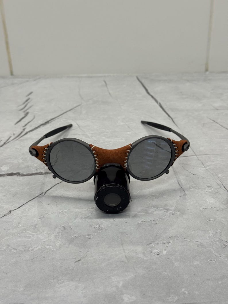 May include: Round sunglasses with dark lenses and a brown leather bridge. The frame features black arms and decorative stitching on the leather. A black cylindrical object is positioned below the glasses.