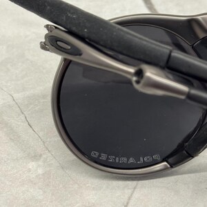 May include: Close-up of dark grey sunglasses with black lenses. The word "POLARISED" is embossed on the frame. The arms are black with a grey metal hinge.