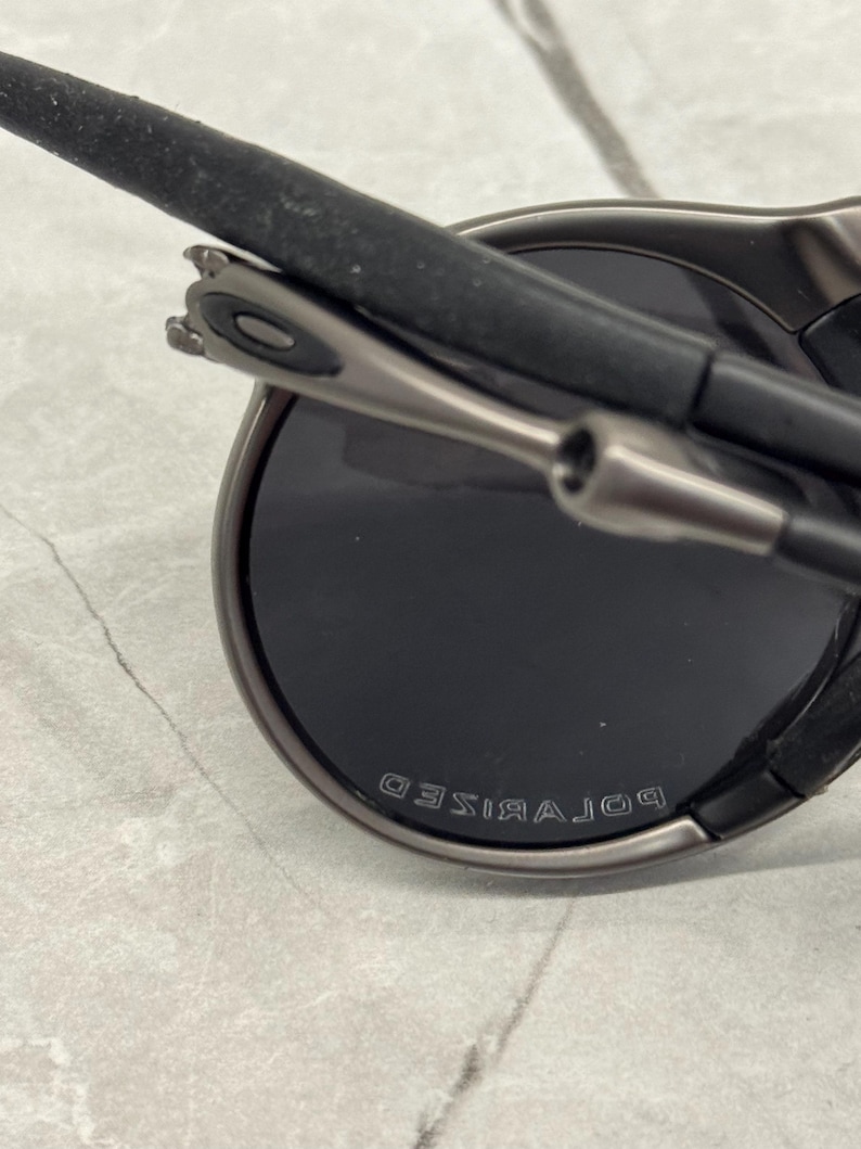 May include: Close-up of dark grey sunglasses with black lenses. The word "POLARISED" is embossed on the frame. The arms are black with a grey metal hinge.