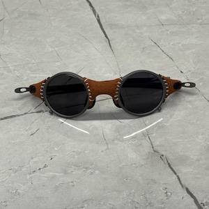 May include: Round sunglasses with dark lenses and a brown leather bridge. The frames are dark gray with visible stitching on the leather. The sunglasses have a unique, vintage aesthetic.