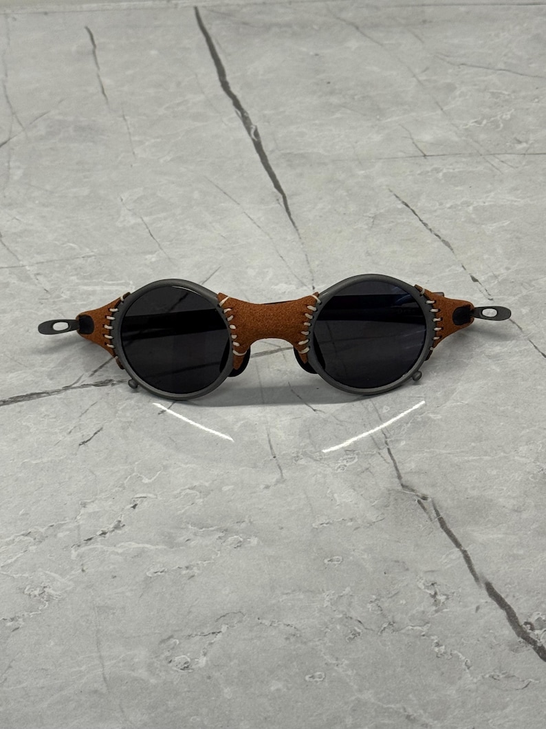 May include: Round sunglasses with dark lenses and a brown leather bridge. The frames are dark gray with visible stitching on the leather. The sunglasses have a unique, vintage aesthetic.