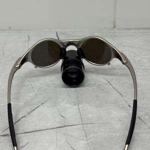 Może przedstawiać: Srebrne okulary przeciwsłoneczne z brązowymi soczewkami. Okulary mają elegancki design z zakrzywionymi zausznikami i czarnymi końc&oacute;wkami. Owalne soczewki kontrastują ze srebrną oprawką. Okulary spoczywają na czarnym stojaku.