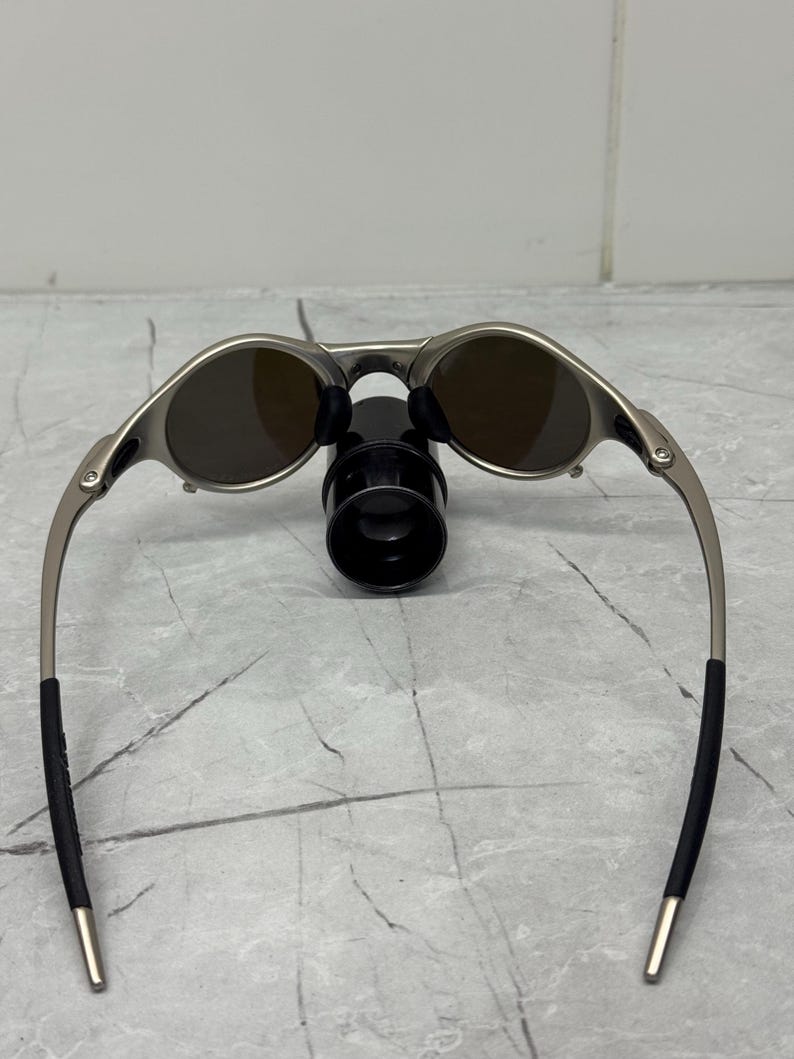 Może przedstawiać: Srebrne okulary przeciwsłoneczne z brązowymi soczewkami. Okulary mają elegancki design z zakrzywionymi zausznikami i czarnymi końc&oacute;wkami. Owalne soczewki kontrastują ze srebrną oprawką. Okulary spoczywają na czarnym stojaku.