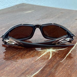 Puede incluir: Primer plano de gafas de sol con montura negra y lentes marrones. Las gafas de sol tienen un acento met&aacute;lico en las patillas y un dise&ntilde;o elegante y moderno. Las lentes son de color marr&oacute;n oscuro y la montura es una combinaci&oacute;n de negro y bronce met&aacute;lico.