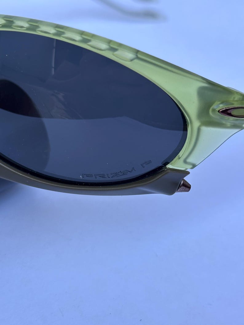 May include: Close-up of a pair of sunglasses with a light green frame and dark lenses. The text "PRIZM" is visible on the lens. The sunglasses have a sleek, modern design.