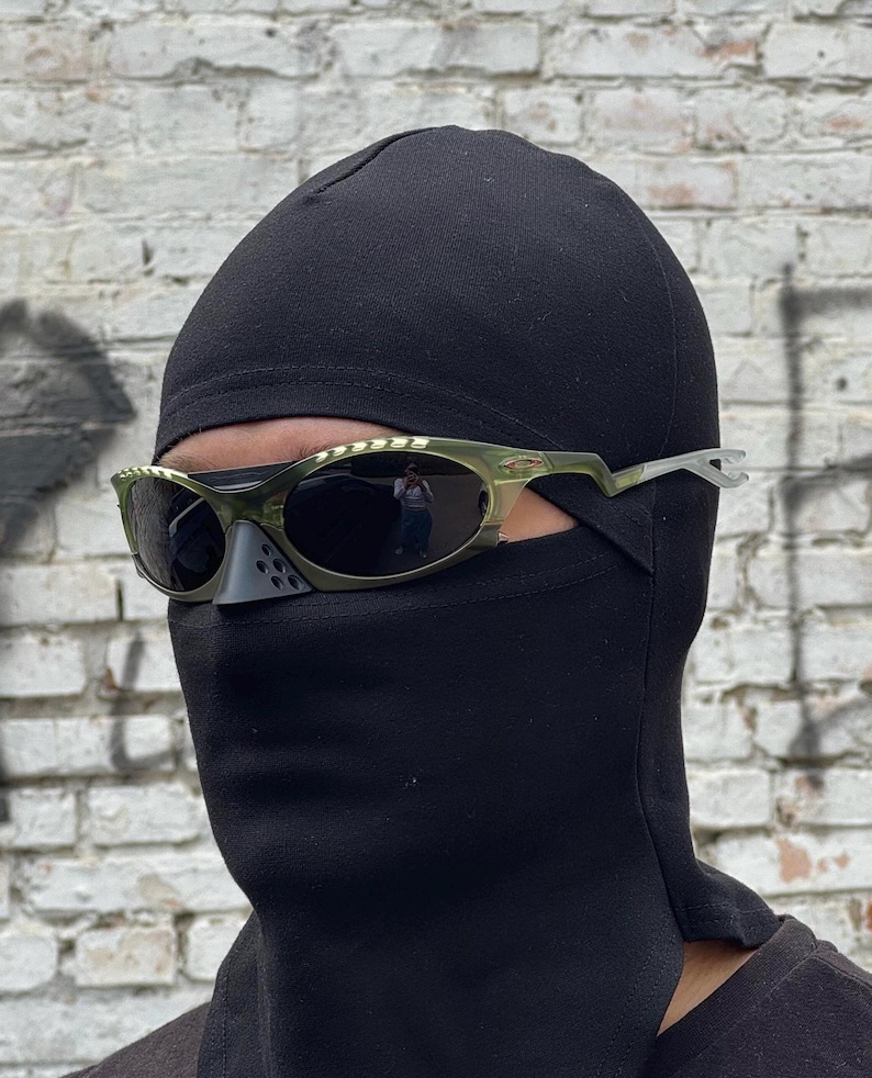 May include: A person wearing a black balaclava and olive green sunglasses with dark lenses. The sunglasses have a unique, angular design. The background is a weathered brick wall.