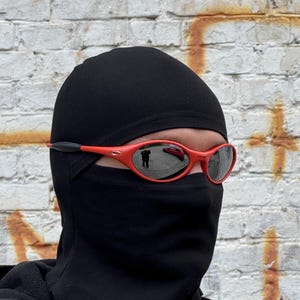 Pu&ograve; includere: Una persona indossa un passamontagna nero e occhiali da sole rossi con lenti riflettenti. Gli occhiali da sole hanno un accento nero sulle aste. Lo sfondo &egrave; un muro di mattoni usurato con vernice arancione e bianca.