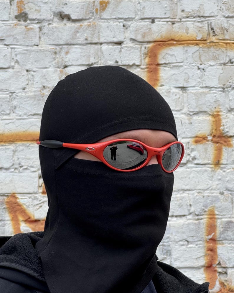 Pu&ograve; includere: Una persona indossa un passamontagna nero e occhiali da sole rossi con lenti riflettenti. Gli occhiali da sole hanno un accento nero sulle aste. Lo sfondo &egrave; un muro di mattoni usurato con vernice arancione e bianca.