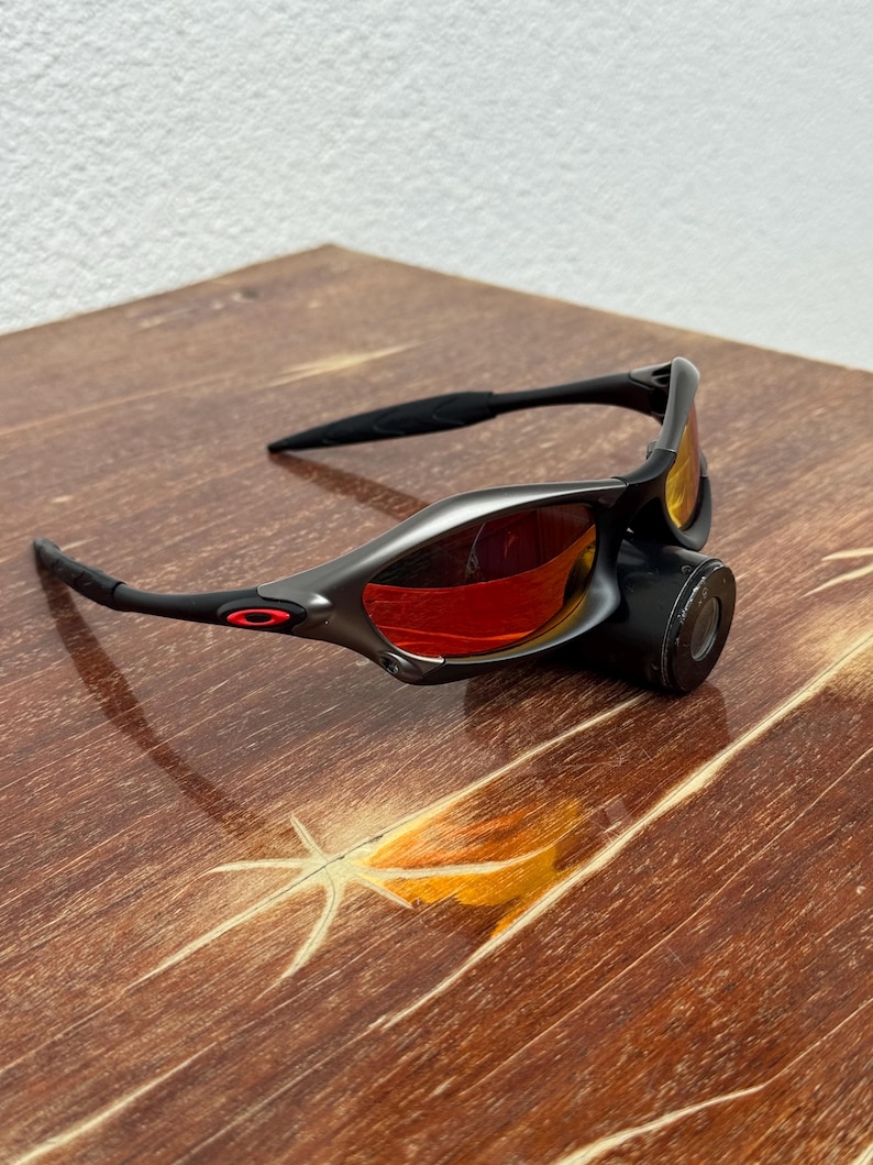 Op de afbeelding: Zwarte en grijze zonnebril met rode glazen. De zonnebril heeft een strak, modern design met een zwart montuur en een rood logo aan de zijkant. De glazen hebben een levendige rode kleur.