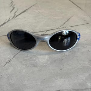 Puede incluir: Gafas de sol plateadas con lentes oscuras. Las gafas tienen un dise&ntilde;o curvo con detalles azules en las patillas. La marca "Oakley" es visible en la montura.