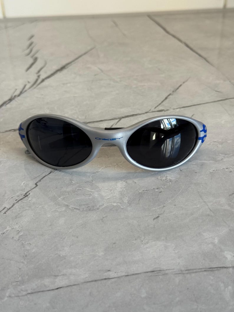 Puede incluir: Gafas de sol plateadas con lentes oscuras. Las gafas tienen un dise&ntilde;o curvo con detalles azules en las patillas. La marca "Oakley" es visible en la montura.