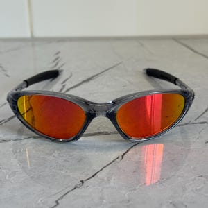 K&ouml;nnte beinhalten: Sportsonnenbrille mit transparentem Rahmen und leuchtend roten Gl&auml;sern. Die Sonnenbrille hat ein schlankes, geschwungenes Design und dunkelgraue B&uuml;gel. Die Gl&auml;ser reflektieren einen hellen Orangeton und bieten einen k&uuml;hnen und sportlichen Look.