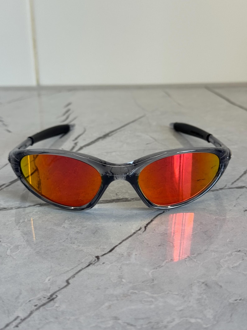 K&ouml;nnte beinhalten: Sportsonnenbrille mit transparentem Rahmen und leuchtend roten Gl&auml;sern. Die Sonnenbrille hat ein schlankes, geschwungenes Design und dunkelgraue B&uuml;gel. Die Gl&auml;ser reflektieren einen hellen Orangeton und bieten einen k&uuml;hnen und sportlichen Look.