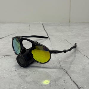 May include: Black sunglasses with yellow and blue lenses. The sunglasses have a unique design with a round lens shape and a black frame. The lenses reflect light, creating a vibrant color effect. The sunglasses are resting on a gray surface.
