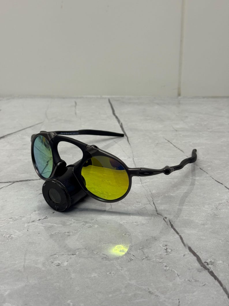 May include: Black sunglasses with yellow and blue lenses. The sunglasses have a unique design with a round lens shape and a black frame. The lenses reflect light, creating a vibrant color effect. The sunglasses are resting on a gray surface.