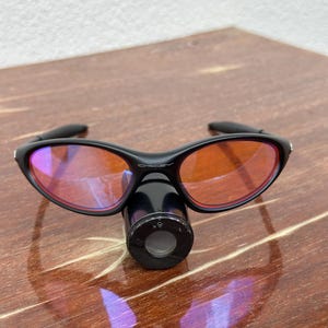 May include: Black sunglasses with orange-tinted lenses and a black cylindrical attachment. The sunglasses have a sleek, modern design. The lenses are oval-shaped and the frame is curved. The attachment has a circular opening with the text "x5".