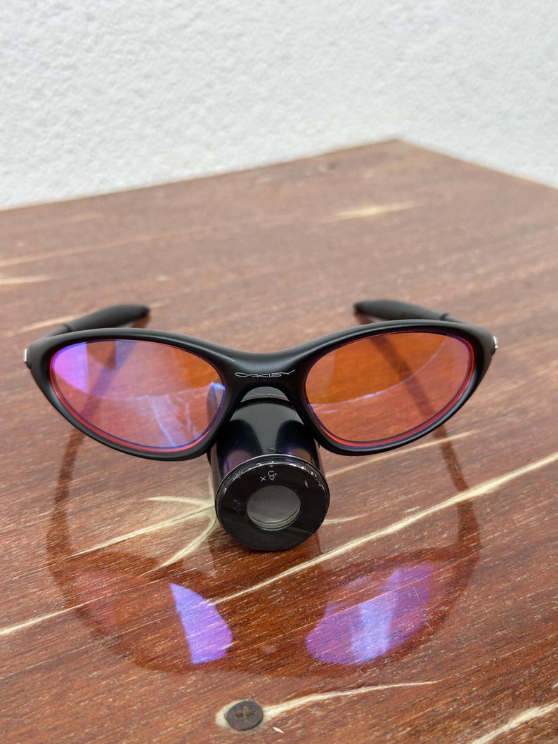 May include: Black sunglasses with orange-tinted lenses and a black cylindrical attachment. The sunglasses have a sleek, modern design. The lenses are oval-shaped and the frame is curved. The attachment has a circular opening with the text "x5".