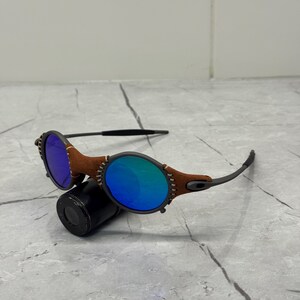 以下が含まれることがあります： 茶色の革のアクセントと青いミラーレンズが付いた丸いサングラス。フレームは濃いグレーで、レンズの周りにステッチされた革のディテールがあります。サングラスは黒い円筒形の物体の上に置かれています。