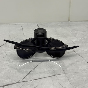 May include: Black sunglasses with a sleek, modern design. The sunglasses feature dark lenses and slender, curved arms with silver accents. The frame is a matte black, and the lenses appear to be circular.
