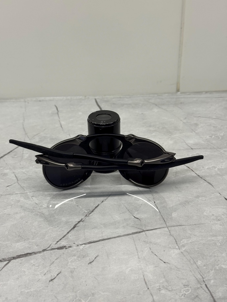 May include: Black sunglasses with a sleek, modern design. The sunglasses feature dark lenses and slender, curved arms with silver accents. The frame is a matte black, and the lenses appear to be circular.
