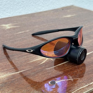 May include: Black sunglasses with orange-tinted lenses and a black cylindrical attachment. The sunglasses have a sleek, modern design with a small logo on the frame. The lenses are a vibrant orange color.