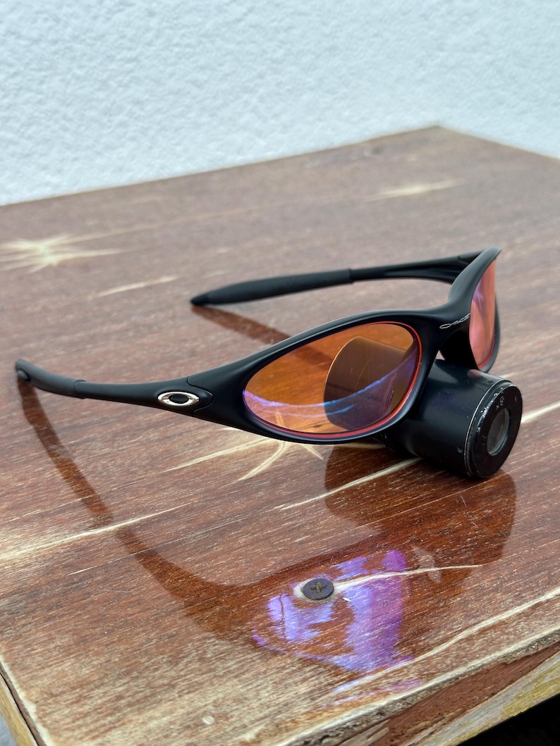 May include: Black sunglasses with orange-tinted lenses and a black cylindrical attachment. The sunglasses have a sleek, modern design with a small logo on the frame. The lenses are a vibrant orange color.