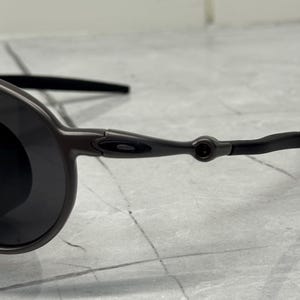 May include: Close-up of a pair of dark gray sunglasses with black lenses. The sunglasses have a sleek, modern design with a thin frame and curved temples. The lenses are round and dark, providing a contrast to the frame.
