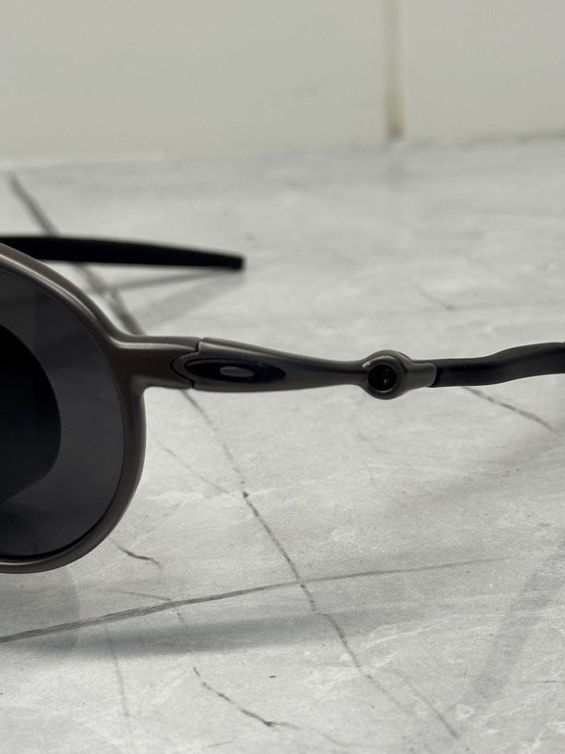 May include: Close-up of a pair of dark gray sunglasses with black lenses. The sunglasses have a sleek, modern design with a thin frame and curved temples. The lenses are round and dark, providing a contrast to the frame.