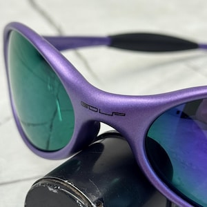 May include: Purple sunglasses with green-tinted lenses. The frame has the word "GOLF" printed on the side. The sunglasses are resting on a black cylindrical object.