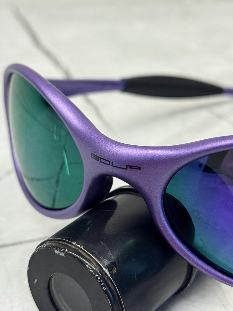 May include: Purple sunglasses with green-tinted lenses. The frame has the word "GOLF" printed on the side. The sunglasses are resting on a black cylindrical object.