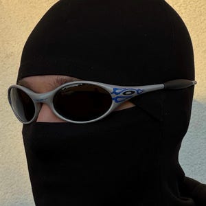 Puede incluir: Gafas de sol plateadas con un dise&ntilde;o de llama azul en las patillas. Las gafas tienen lentes oscuras y se llevan con un pasamonta&ntilde;as negro. El pasamonta&ntilde;as cubre la cabeza y el cuello, dejando solo los ojos visibles.