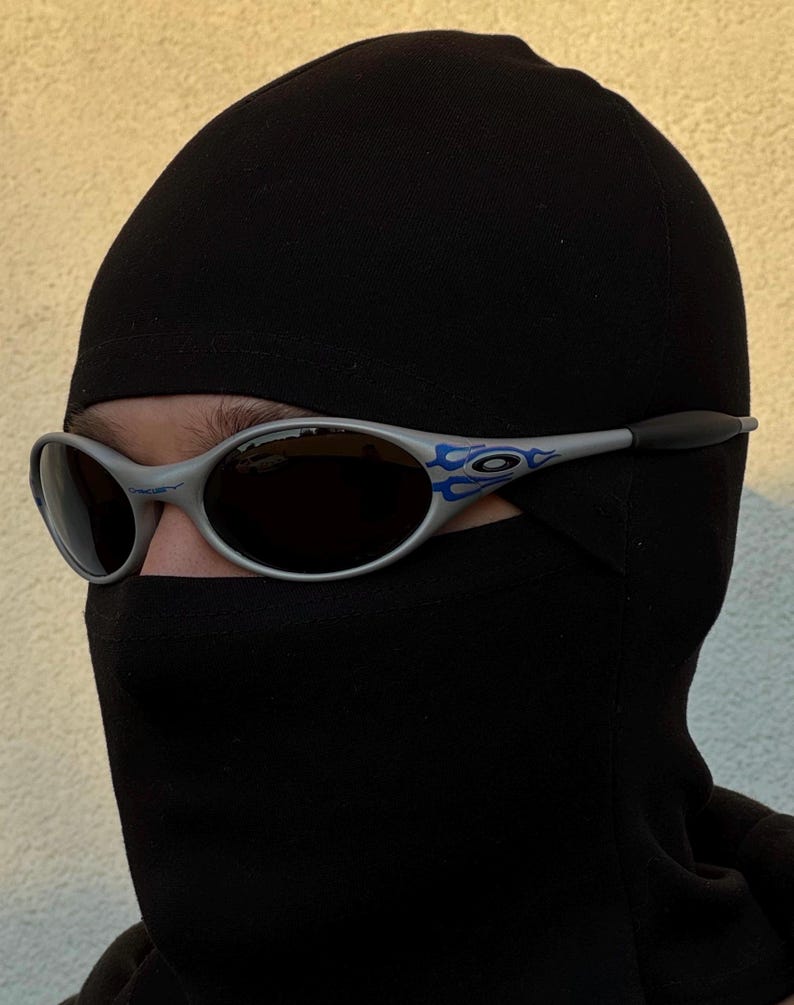 Puede incluir: Gafas de sol plateadas con un dise&ntilde;o de llama azul en las patillas. Las gafas tienen lentes oscuras y se llevan con un pasamonta&ntilde;as negro. El pasamonta&ntilde;as cubre la cabeza y el cuello, dejando solo los ojos visibles.