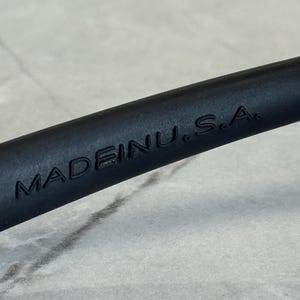 K&ouml;nnte beinhalten: Nahaufnahme eines schwarzen Objekts mit dem gepr&auml;gten Text "MADE INU. S.A.". Das Objekt scheint Teil eines gr&ouml;&szlig;eren Gegenstands zu sein, m&ouml;glicherweise ein Werkzeug oder Zubeh&ouml;r. Der Hintergrund ist eine hellgraue, strukturierte Oberfl&auml;che.