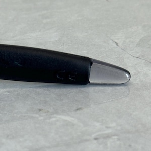 K&ouml;nnte beinhalten: Nahaufnahme eines schwarzen Brillenb&uuml;gels mit einer klaren, abgerundeten Spitze. Der B&uuml;gel hat eine matte Oberfl&auml;che und der Text "CE" ist sichtbar. Der Hintergrund ist eine hellgraue, strukturierte Oberfl&auml;che.