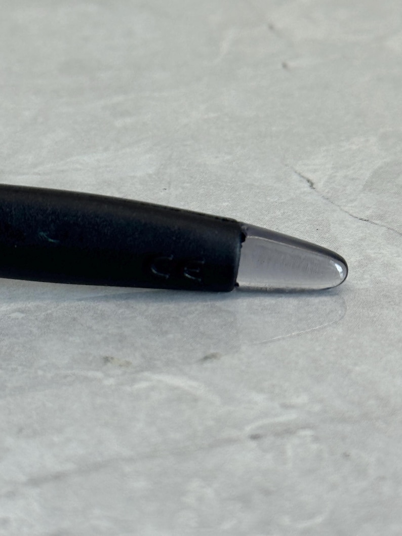 K&ouml;nnte beinhalten: Nahaufnahme eines schwarzen Brillenb&uuml;gels mit einer klaren, abgerundeten Spitze. Der B&uuml;gel hat eine matte Oberfl&auml;che und der Text "CE" ist sichtbar. Der Hintergrund ist eine hellgraue, strukturierte Oberfl&auml;che.