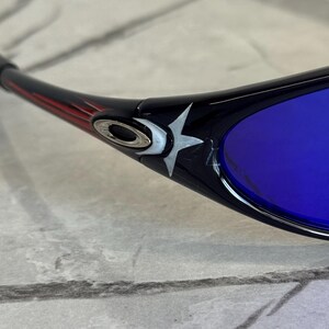 May include: Black sunglasses with blue lenses. The frame features a red and black design with a silver logo and a white star emblem. The lenses are a vibrant blue color, offering a striking contrast.