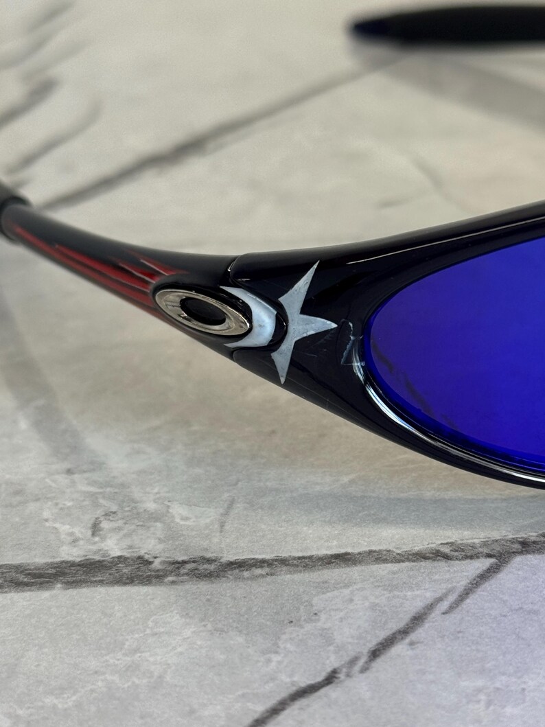 May include: Black sunglasses with blue lenses. The frame features a red and black design with a silver logo and a white star emblem. The lenses are a vibrant blue color, offering a striking contrast.