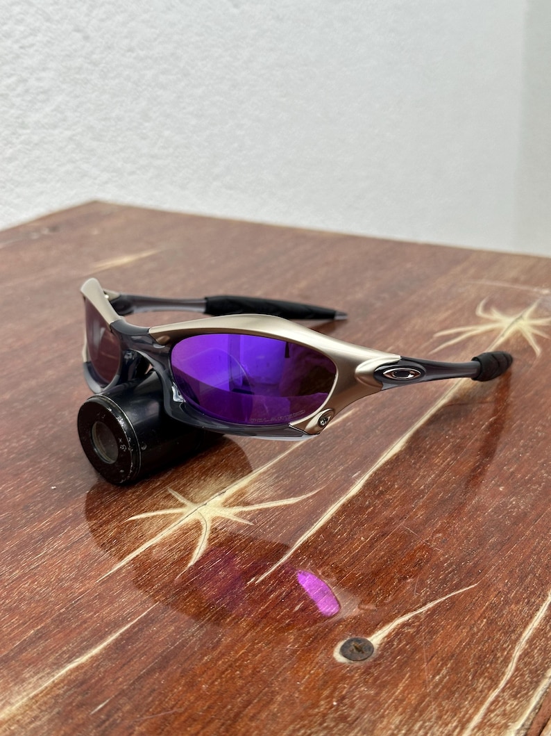 May include: Silver and black sunglasses with purple lenses. The sunglasses are on a wooden surface.